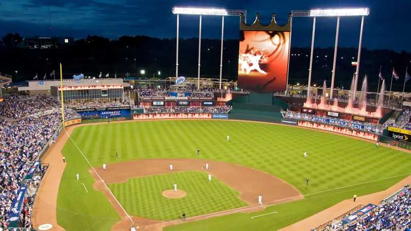 Kauffman Stadium at dusk during a night game, with the crown-topped scoreboard and the outfield fountains lit