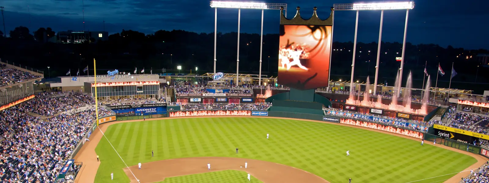 Kauffman Stadium at dusk during a night game, with the crown-topped scoreboard and the outfield fountains lit